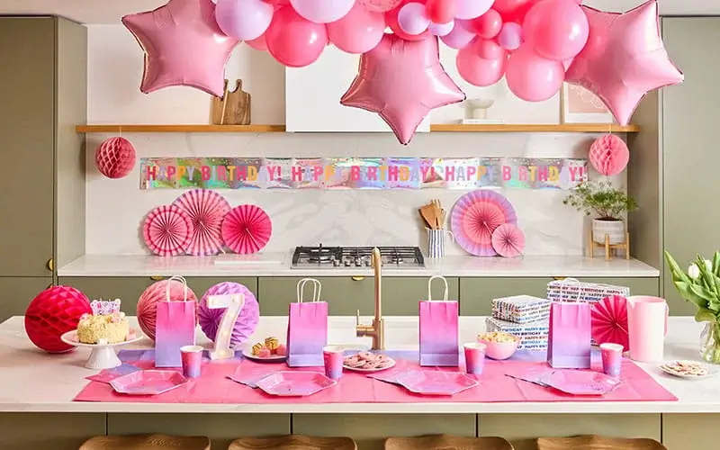 Pink birthday party table with balloon garland, star balloons, a lit number 7 decoration, gift bags, party tableware, and birthday decorations.